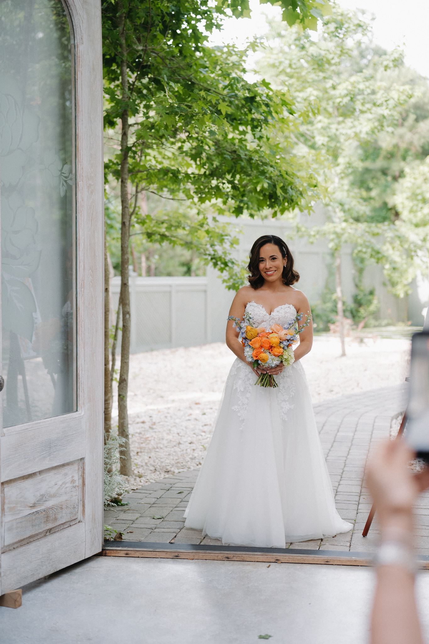 Bride in white strapless ball gown holding vibrant orange bouquet stands in doorway surrounded by lush green trees.