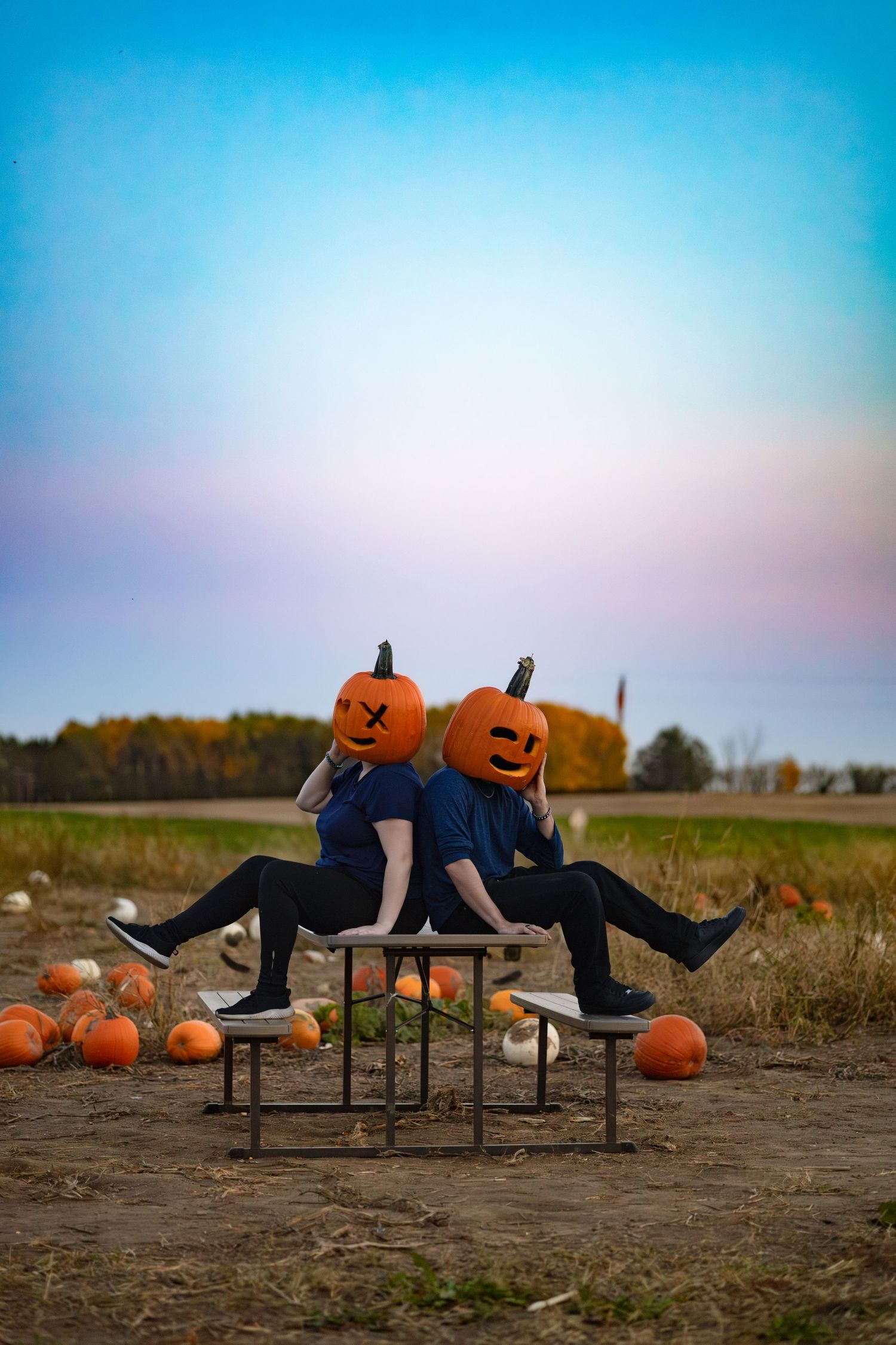 Two people wearing jack-o-lantern pumpkin heads sit back-to-back on a bench in a pumpkin patch at dusk.
