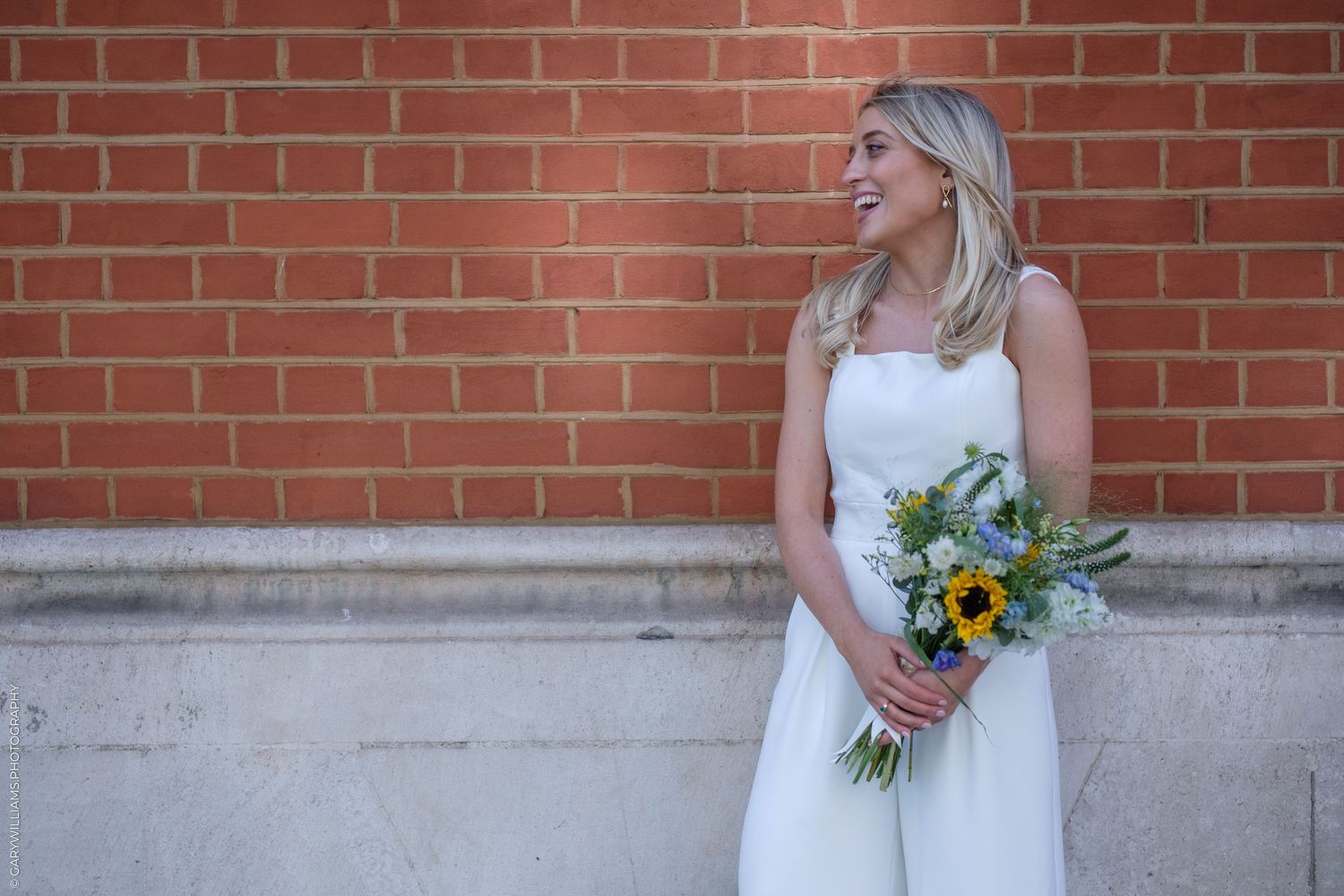 Smiling bride in white dress holds sunflower bouquet standing against a red brick wall outdoors.