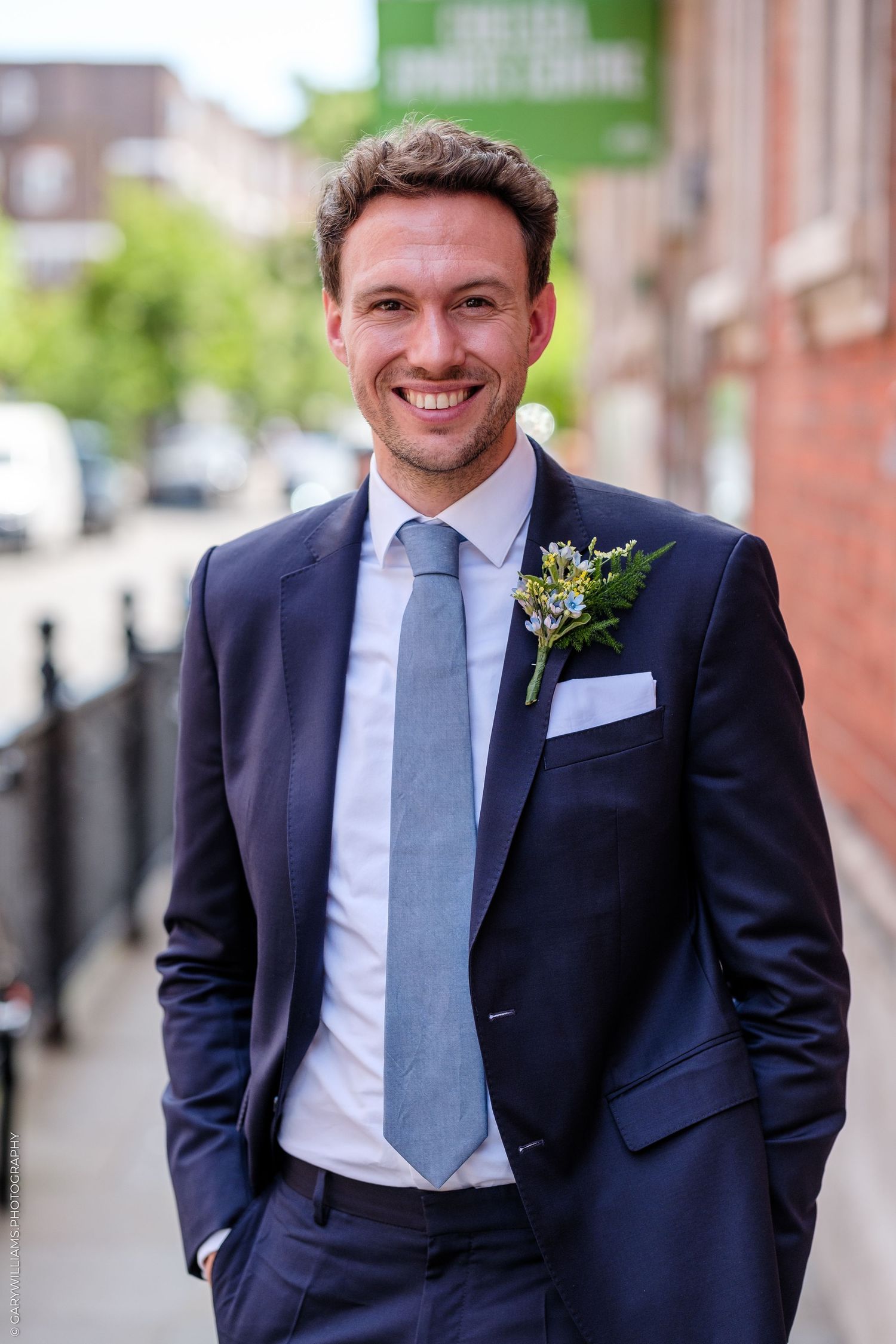 Smiling groom in navy suit with blue tie and floral boutonniere standing on a sunny city street.