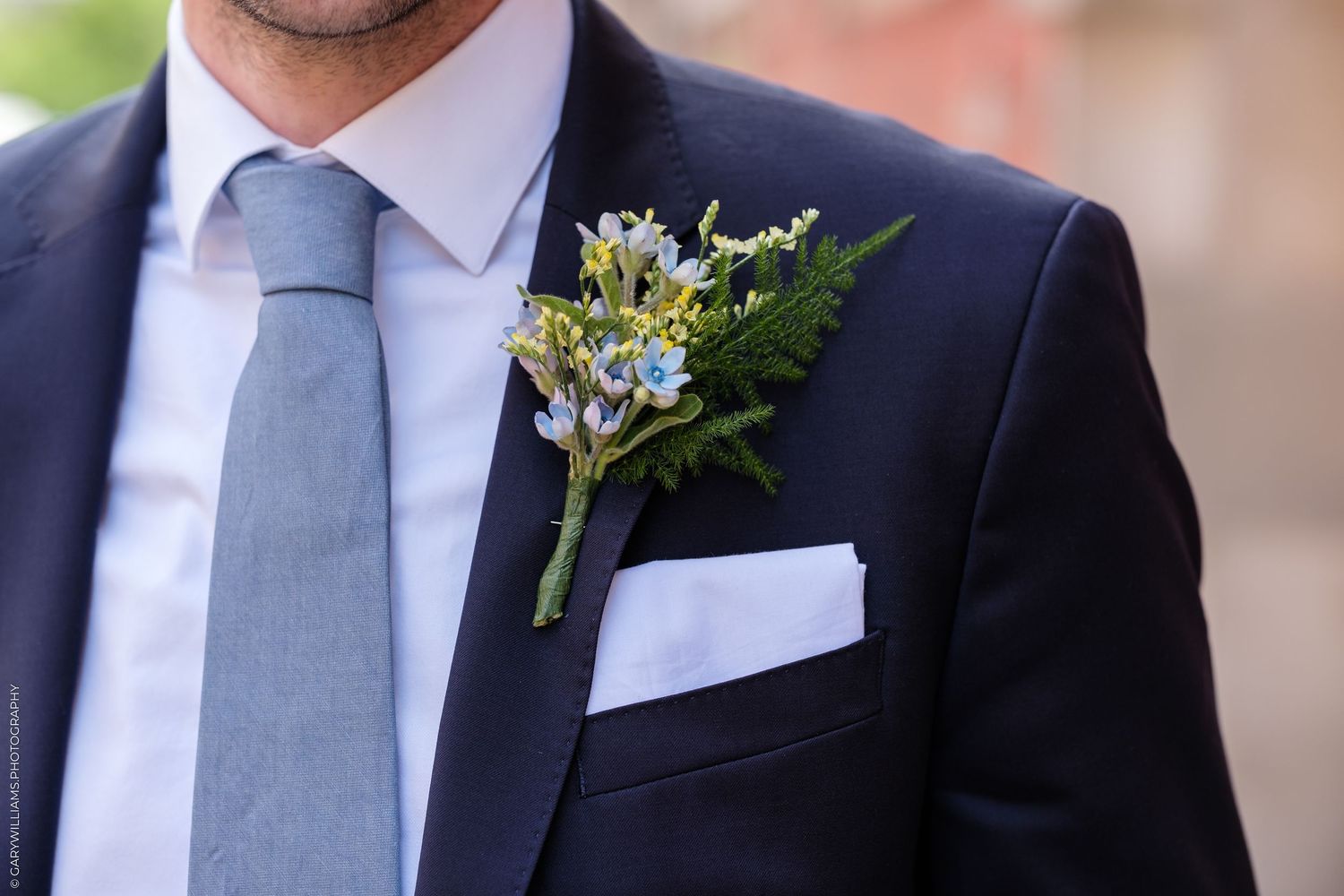 Close-up of a groom in a navy suit with a blue tie, white pocket square, and floral boutonniere with greenery.