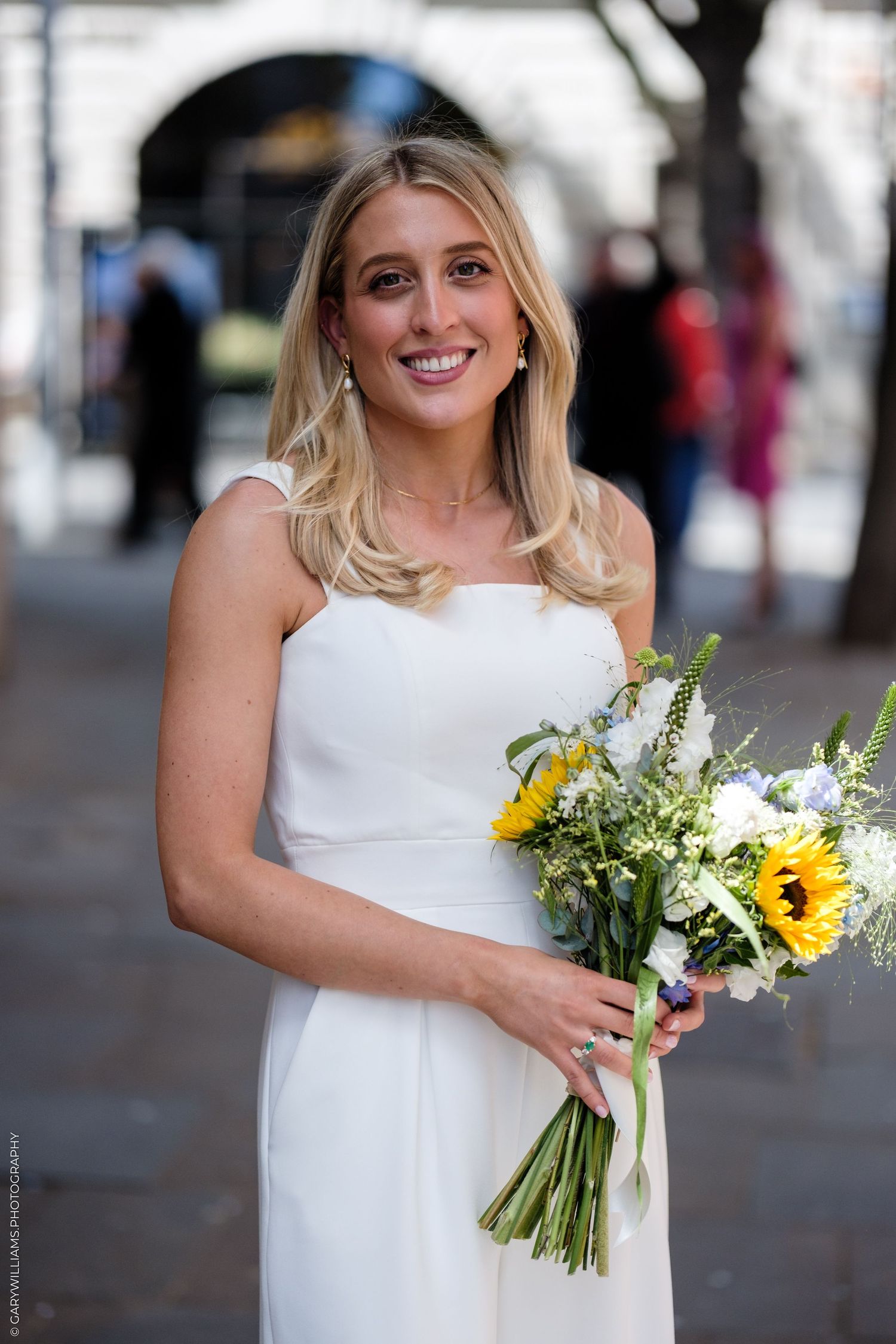 Radiant bride in white dress holding sunflower and wildflower bouquet smiling outdoors on wedding day.