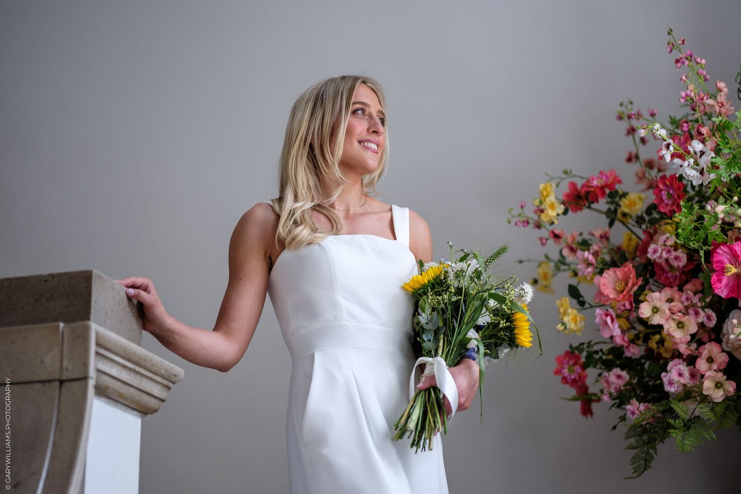 Blonde bride in white gown holding yellow floral bouquet stands near large pink flower arrangement in studio.