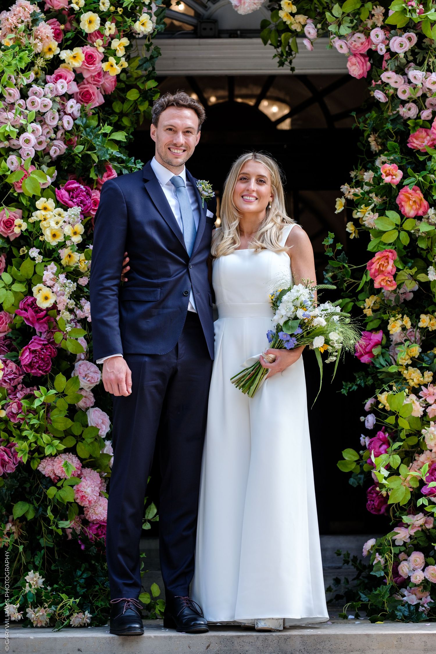 Bride and groom posing in front of a colorful floral arch, bride holding a bouquet, groom in a navy suit.