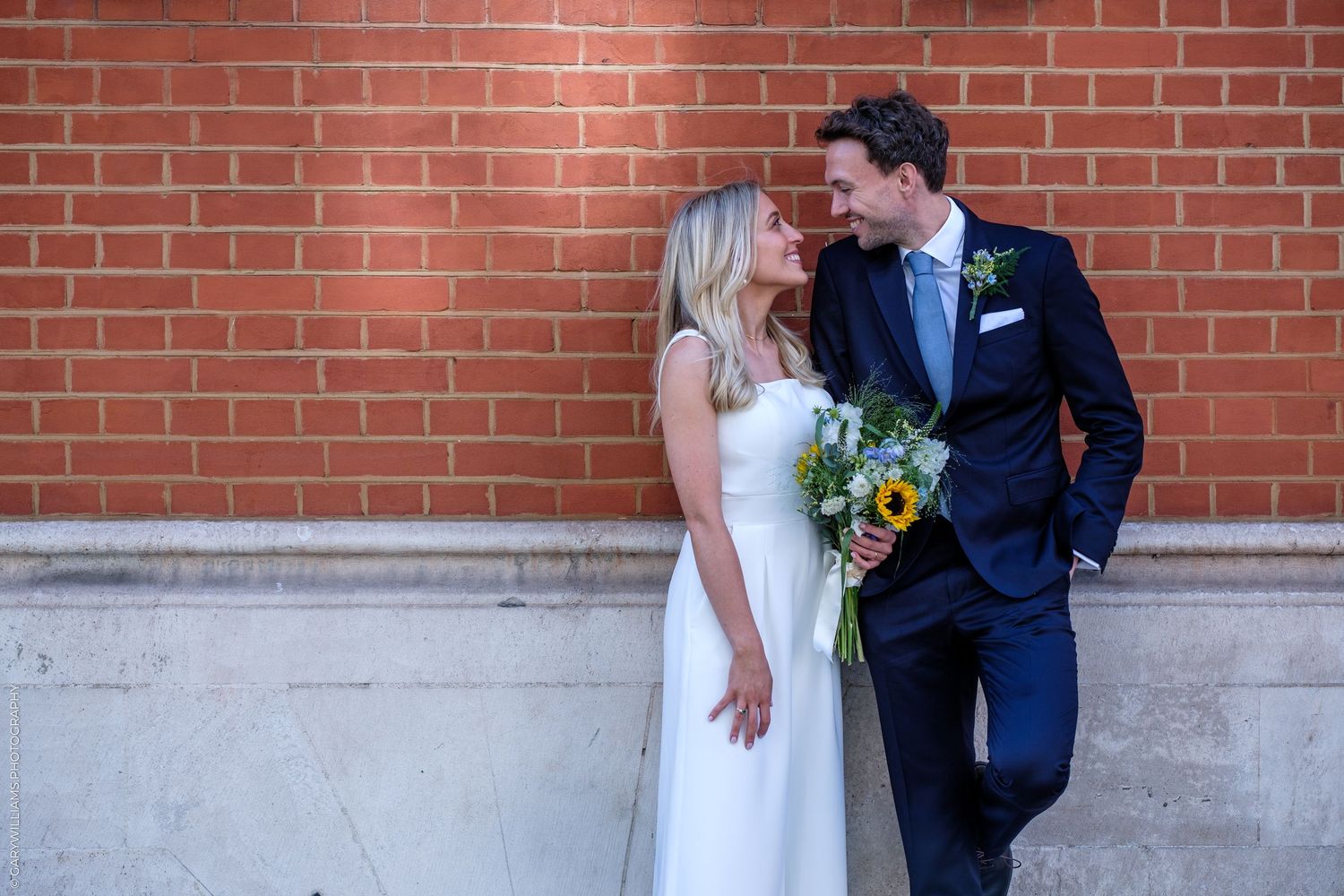 Bride and groom smiling at each other against brick wall, bride holding sunflower bouquet, groom in navy suit.