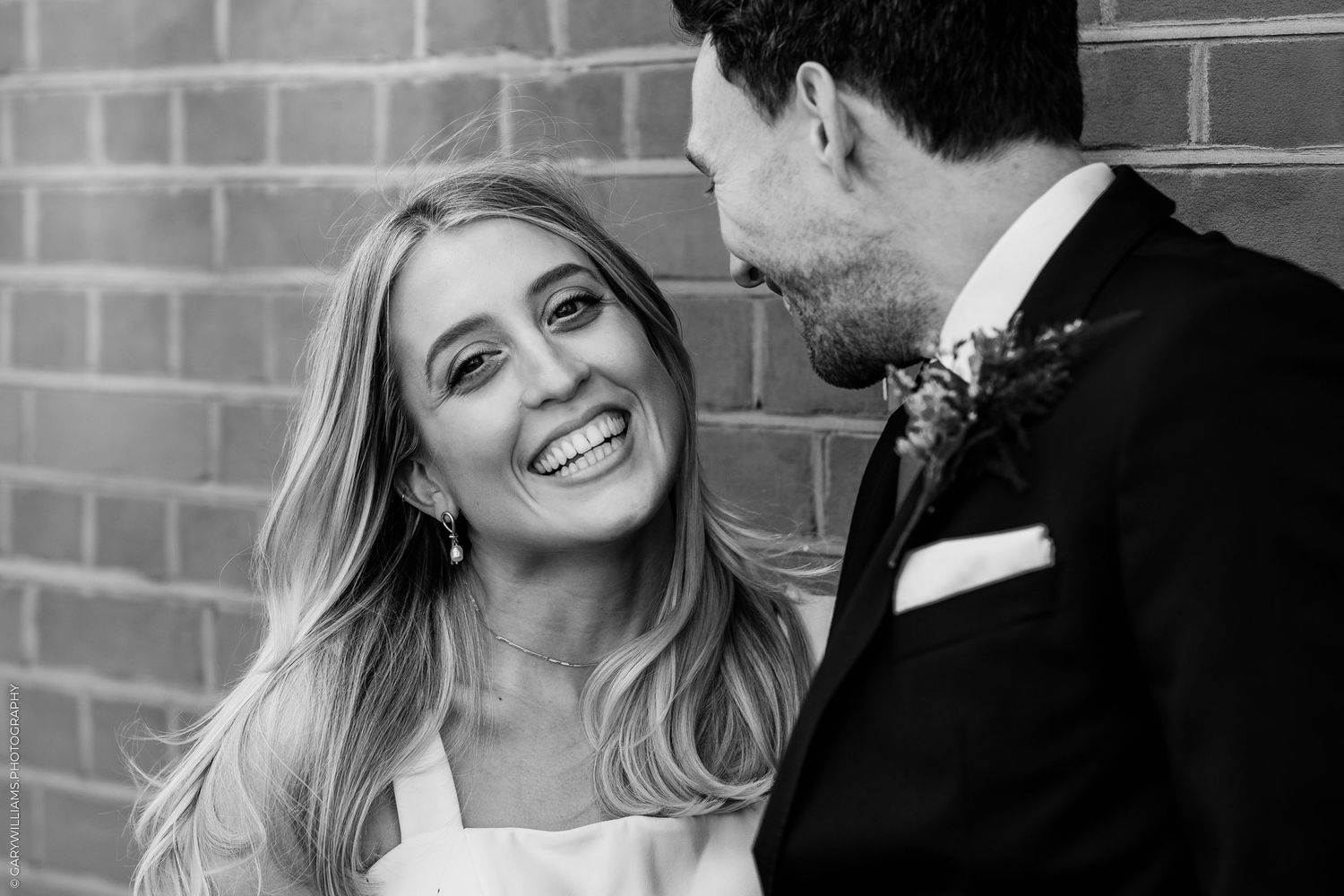 Black and white wedding photo of smiling bride looking up at groom in suit with boutonniere against brick wall.