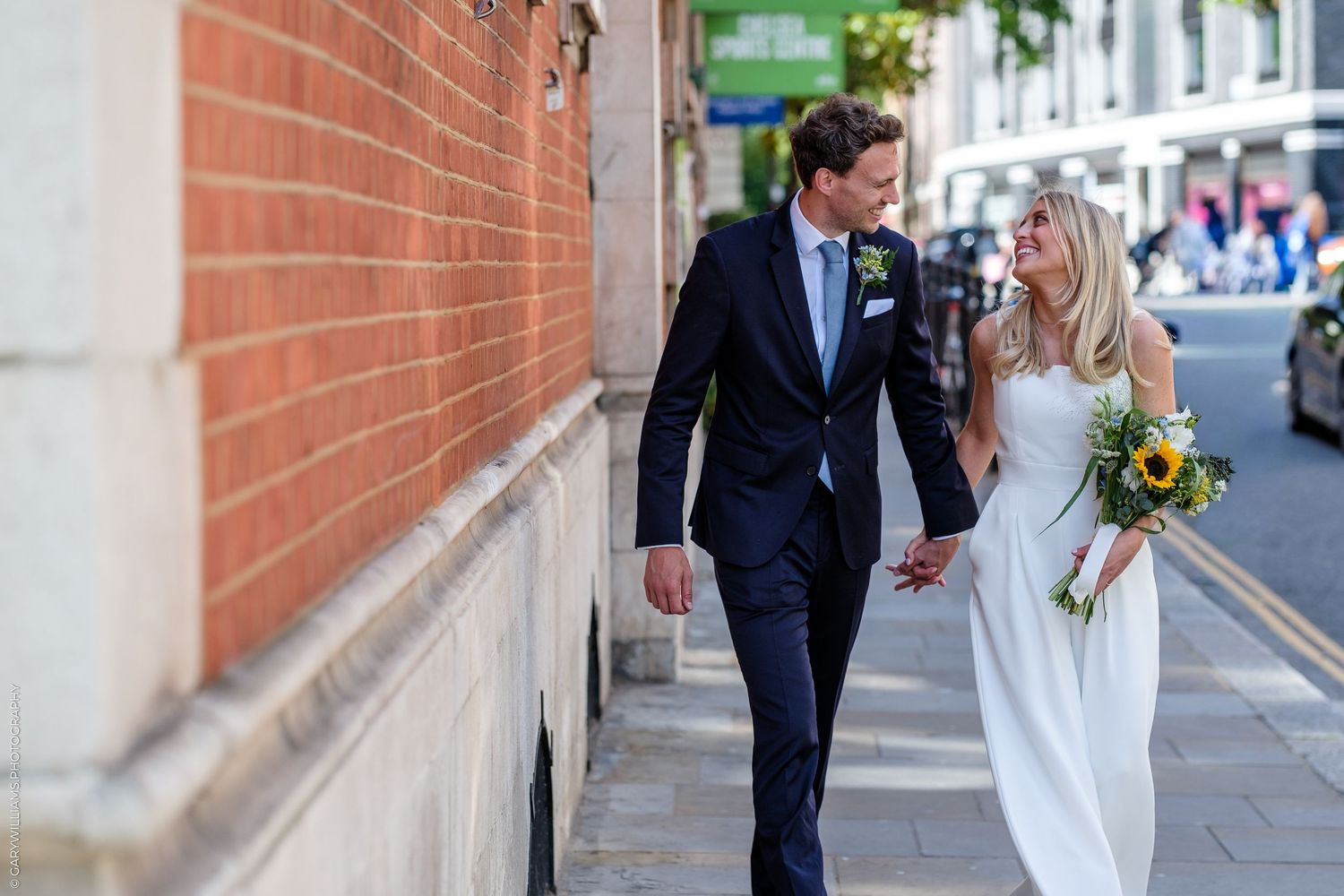 Newlywed couple walking hand in hand on a sunny city street, bride holding sunflower bouquet in white gown.