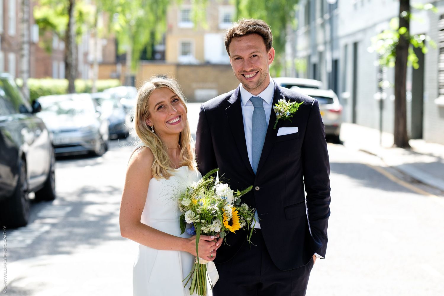 Smiling bride and groom pose on a sunny street, bride holding yellow and white bouquet, groom in navy suit with boutonniere.