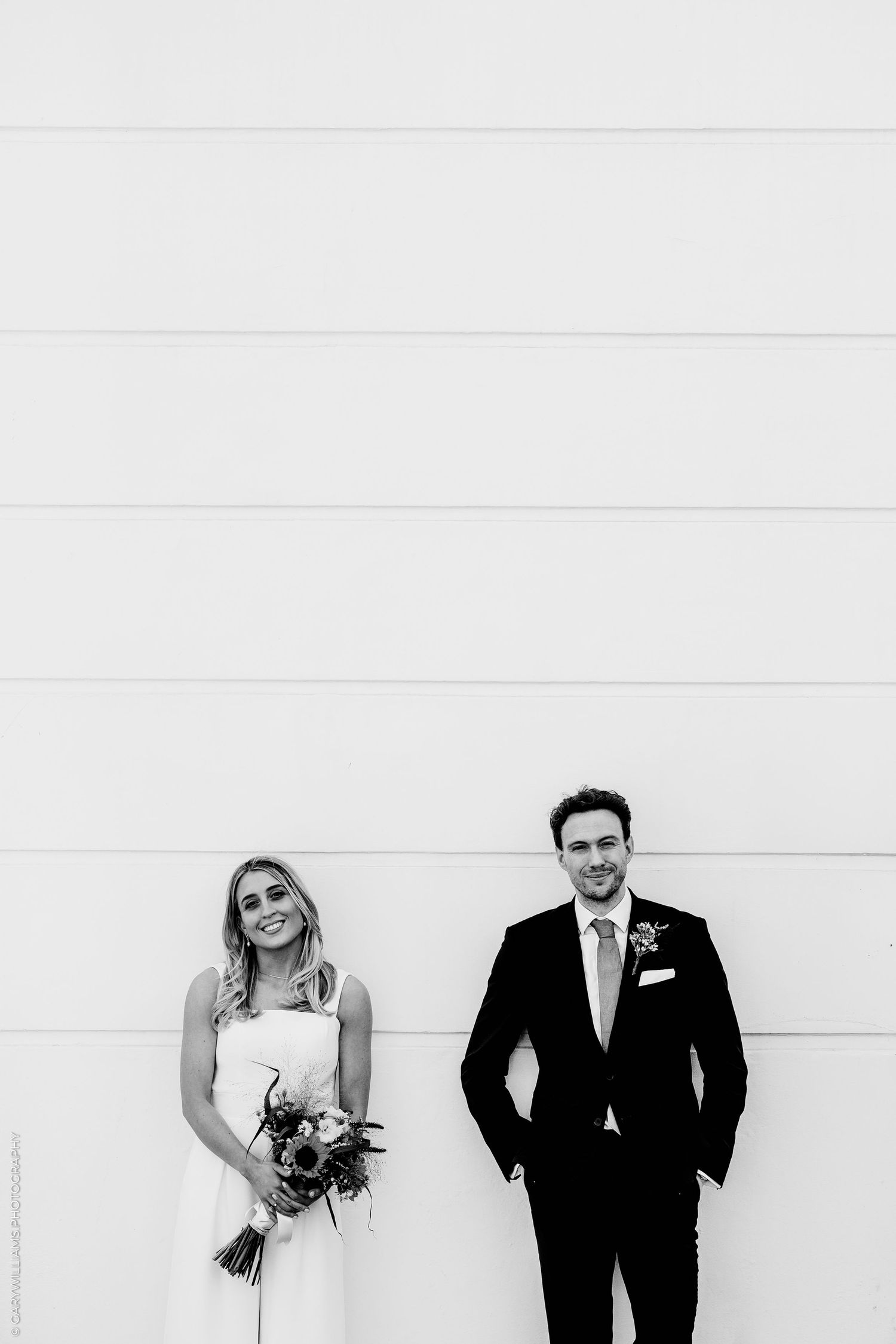 Black and white photo of a smiling bride holding a bouquet and groom in a suit standing against a white wall.