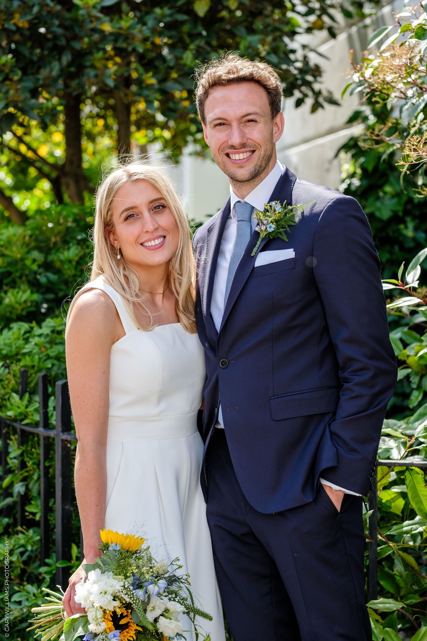 Smiling bride and groom pose outdoors in wedding attire, she holds sunflower bouquet, he wears navy suit with boutonniere.