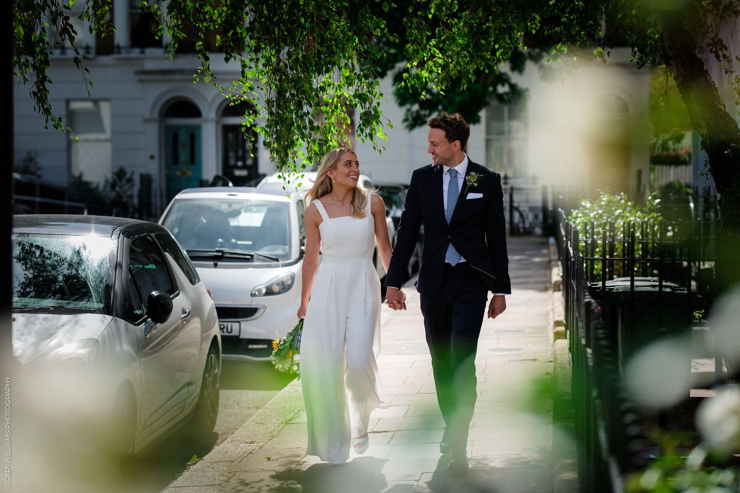 Couple holding hands walking on a sunny London street, bride in white dress and groom in navy suit.