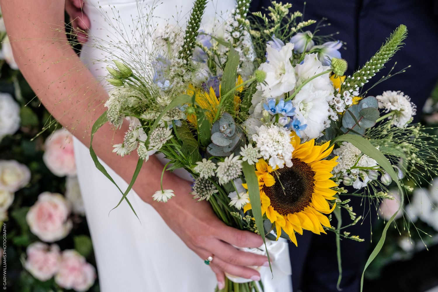 Bride holding a rustic wedding bouquet featuring a sunflower, white blooms, blue flowers, and greenery at an outdoor ceremony.