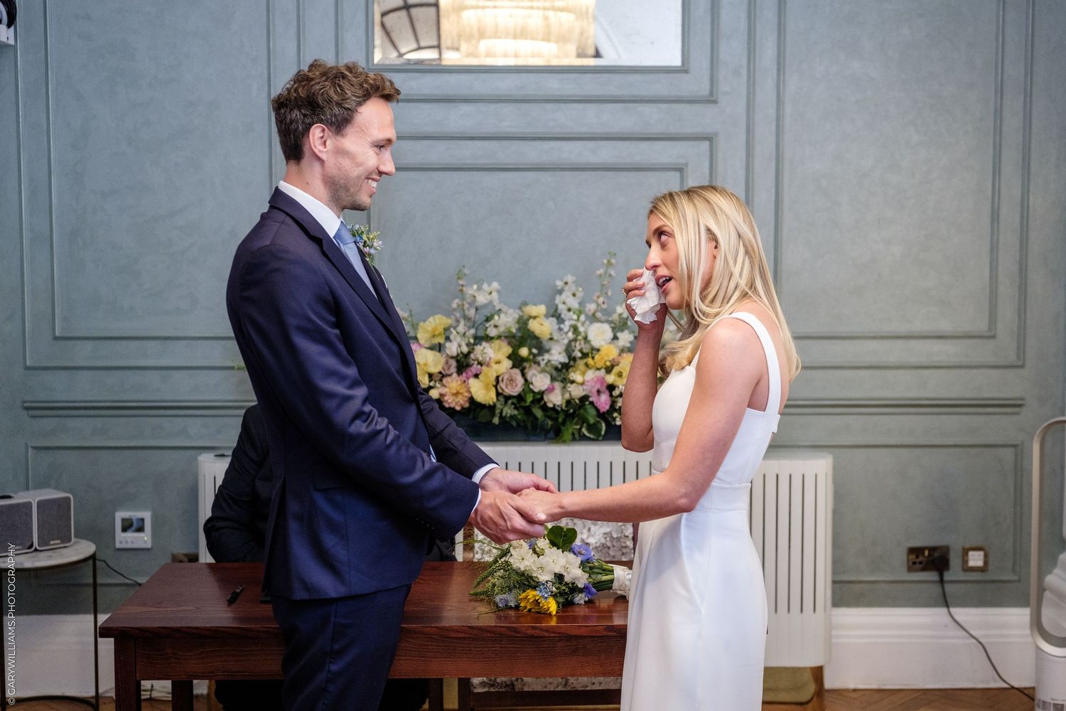 Emotional bride and groom holding hands during intimate wedding ceremony in an elegant room with floral arrangement.