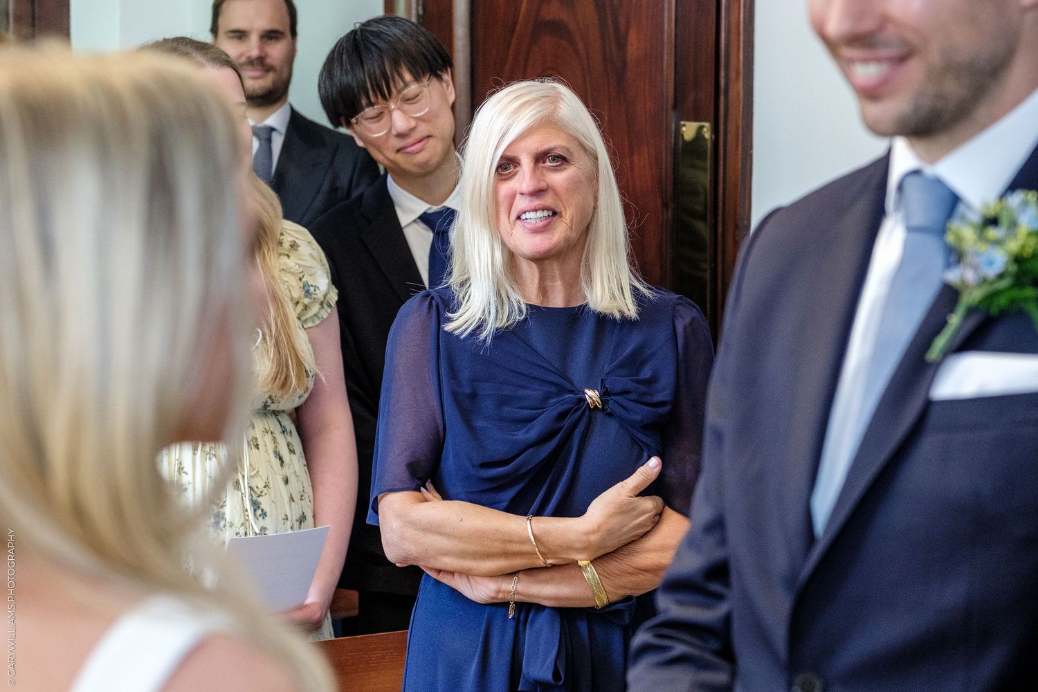 Woman in navy dress smiles warmly at a wedding reception with guests in formal attire in background.
