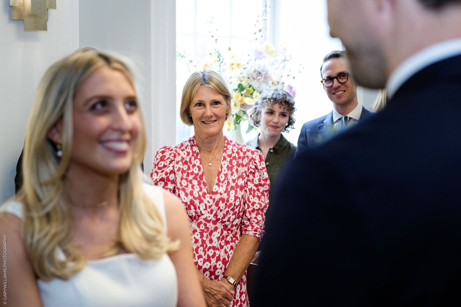 Guests smiling warmly at a wedding ceremony, woman in red floral dress standing in background.