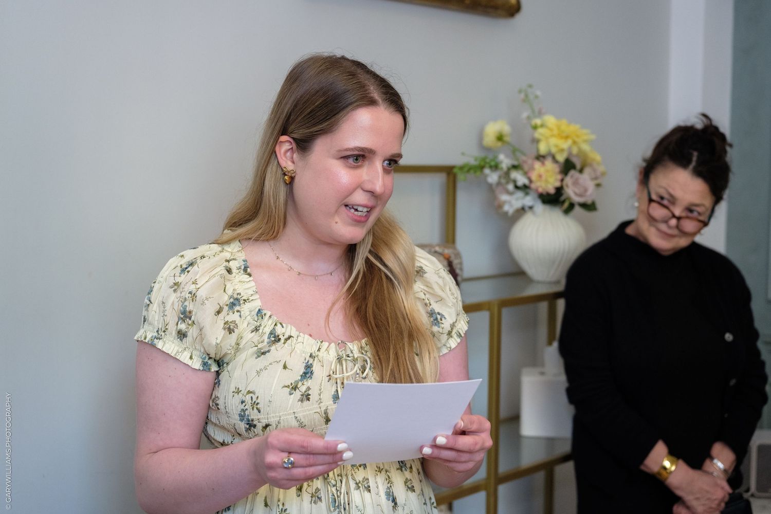 Young woman in floral dress reads from paper at indoor event, with another woman standing in background.