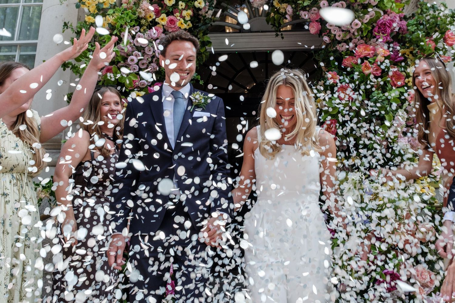 Bride and groom exit wedding venue through confetti shower under floral arch with bridesmaids celebrating.