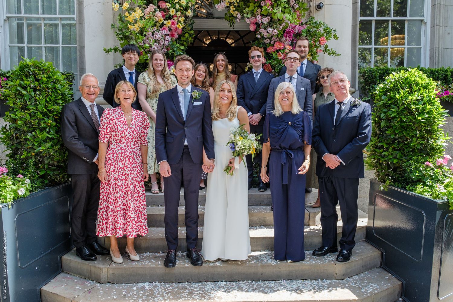 Wedding party posing on steps outside a floral-decorated venue, bride in white gown with bouquet, groom in navy suit.