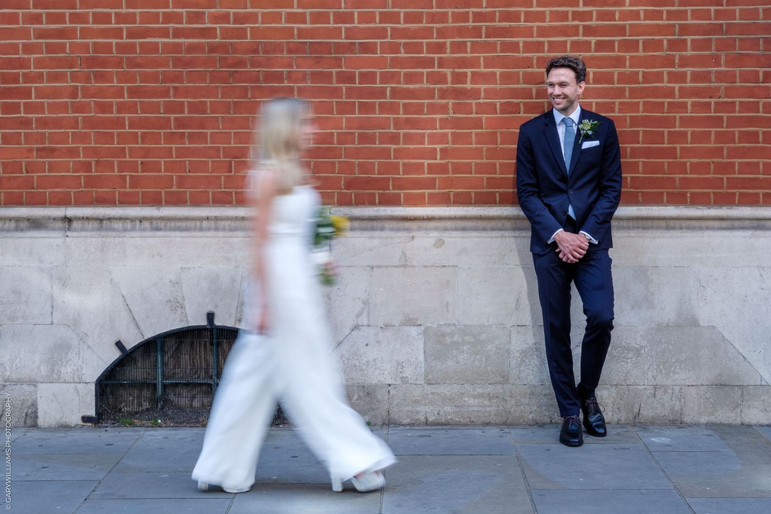 Groom in navy suit smiles at bride in white dress walking past brick wall outdoor wedding photo.