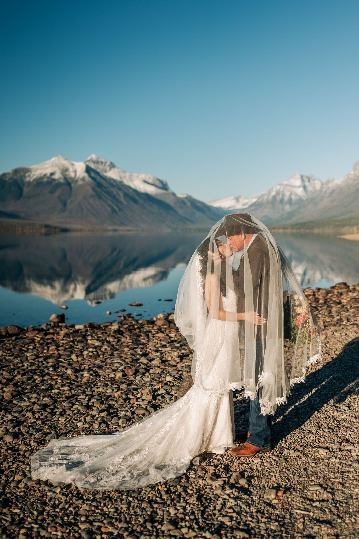 Bride and groom share a kiss under a flowing veil by a serene mountain lake with snow-capped peaks reflecting in calm waters.