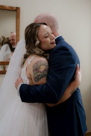 Bride in lace gown with tattoos shares emotional hug with groom in navy suit during wedding day first look.
