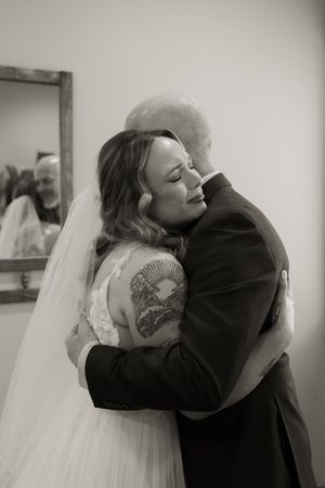 Emotional black and white photo of a bride in lace wedding dress and veil sharing a tender hug with a bald man indoors.