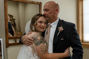 Smiling bride with tattoos in lace wedding dress shares a warm embrace with a bald man in a navy suit indoors.
