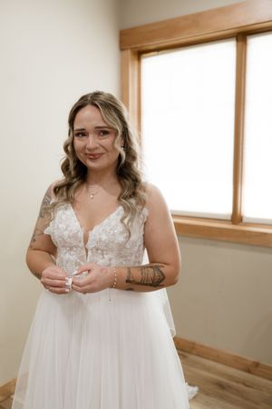Beautiful bride in lace wedding dress with tattoos poses by window before ceremony.