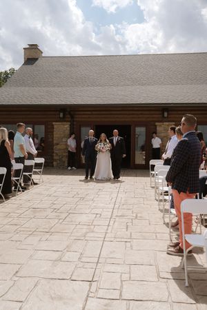 Bride walks down aisle escorted by two men at outdoor wedding ceremony on stone patio.