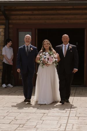 Bride in white gown holding pink bouquet escorted by two men outside rustic venue.