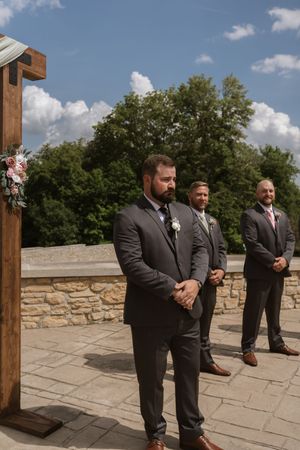 Groom and groomsmen standing at outdoor wedding altar on stone patio with blue sky and trees.
