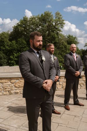 Groom in dark suit with boutonniere stands with groomsmen outdoors at stone wedding venue under blue sky.