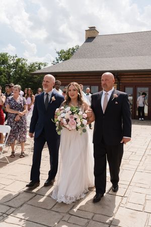 Bride walks down outdoor wedding aisle escorted by two men in navy suits, holding pink and white bouquet.