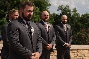 Groomsmen in charcoal suits with boutonnieres standing solemnly during outdoor wedding ceremony.