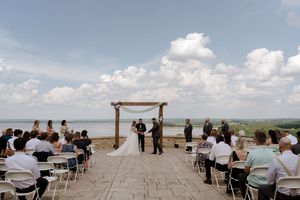 Outdoor lakeside wedding ceremony under wooden arch with bride, groom, officiant, and guests seated in white chairs.