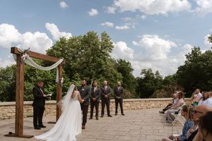 Outdoor wedding ceremony on stone terrace with wooden arch, groomsmen, and seated guests.