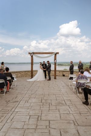 Outdoor wedding ceremony on stone patio overlooking lake with wooden arch draped in white fabric and seated guests.