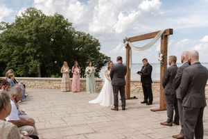 Outdoor wedding ceremony on stone patio with wooden arch, bride and groom exchanging vows with bridesmaids and guests.