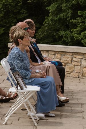 Elegantly dressed wedding guests seated on white folding chairs at outdoor stone terrace ceremony.