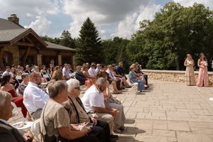 Wedding guests seated outdoors on stone patio near rustic log cabin surrounded by lush green trees.
