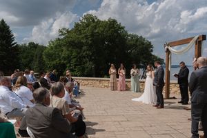Outdoor wedding ceremony on stone patio with guests seated, bridal party at floral arch overlooking trees.