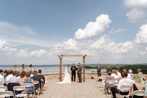 Wide shot of outdoor lakeside wedding ceremony with wooden arch, seated guests, and dramatic cloudy blue sky.