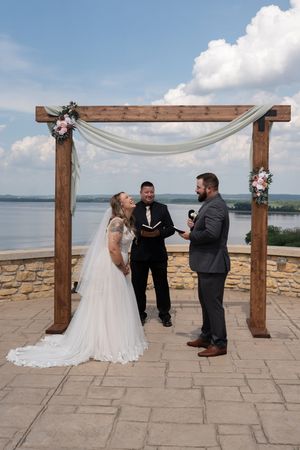 Bride and groom exchange vows under rustic wooden arch with white draping and lake view backdrop.