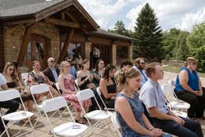 Wedding guests seated in white folding chairs on outdoor patio beside a rustic log cabin venue.