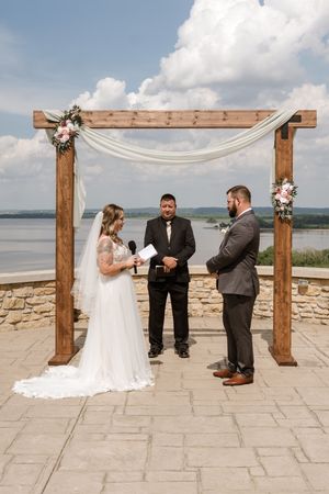 Bride and groom exchange vows under a rustic wooden arch with floral decor, overlooking a river on a sunny day.