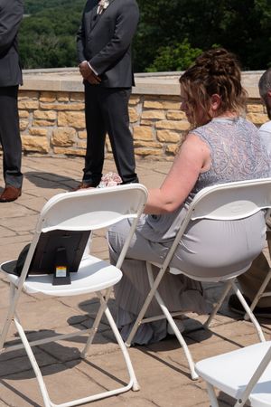 Wedding guest seated in white folding chair holding program during outdoor stone terrace ceremony.