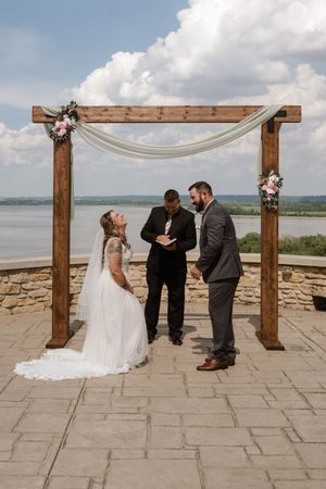 Bride and groom exchange vows under a wooden arch with floral decor overlooking a river.