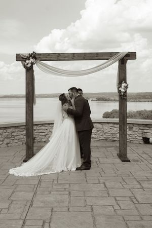 Bride and groom share first kiss under rustic wooden arch with lake view in black and white.
