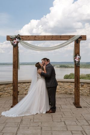 Bride and groom share first kiss under rustic wooden arch with floral accents overlooking scenic lake.
