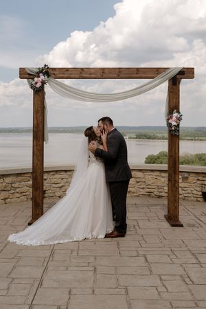Newlyweds kiss beneath draped wooden wedding arch adorned with pink flowers overlooking a scenic river.